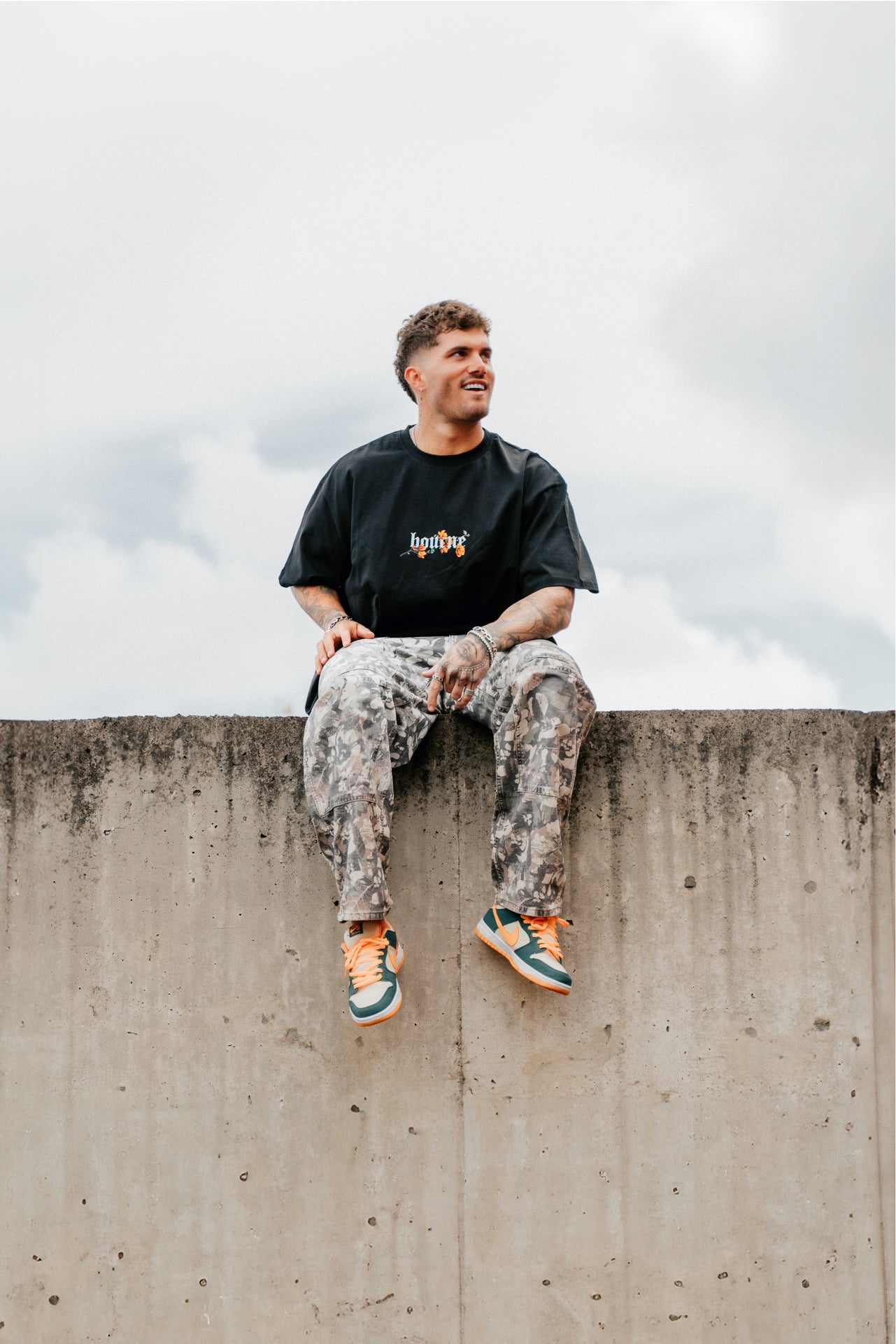 male model sitting on a concrete wall, smiling, wearing a flower streetwear t-shirt, and cargo pants