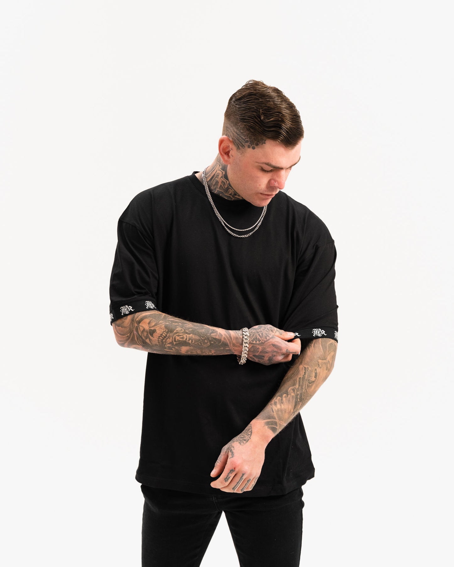 male model with tattoos and chains wearing black t-shirt and black ripped jeans, white background