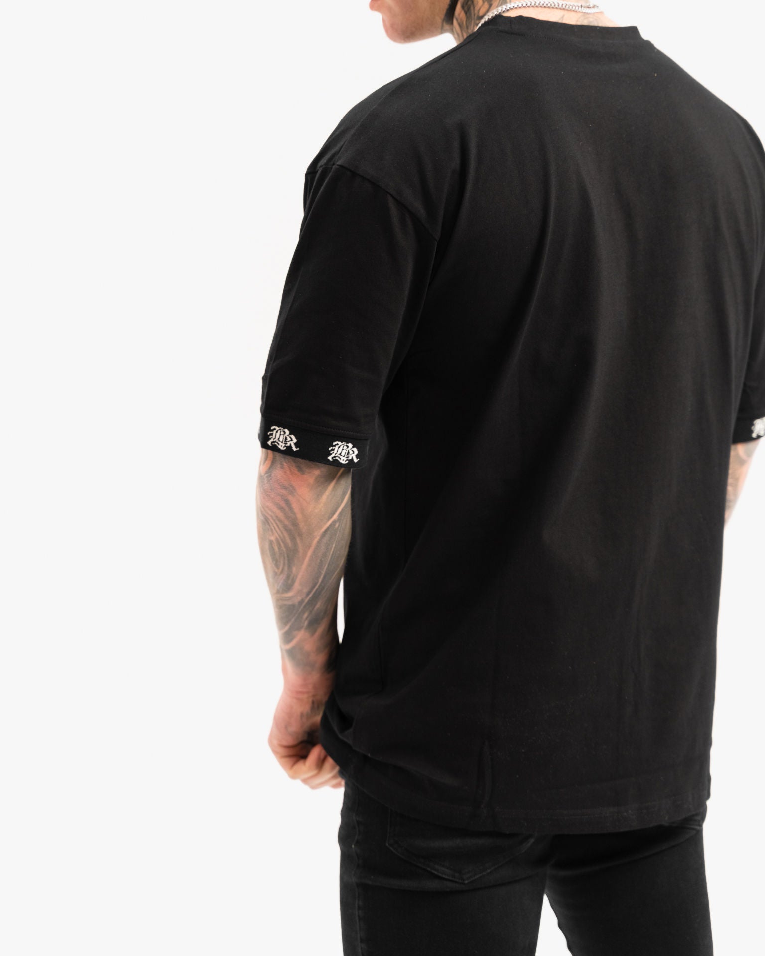 male model with tattoos and chains wearing black t-shirt and black ripped jeans, white background