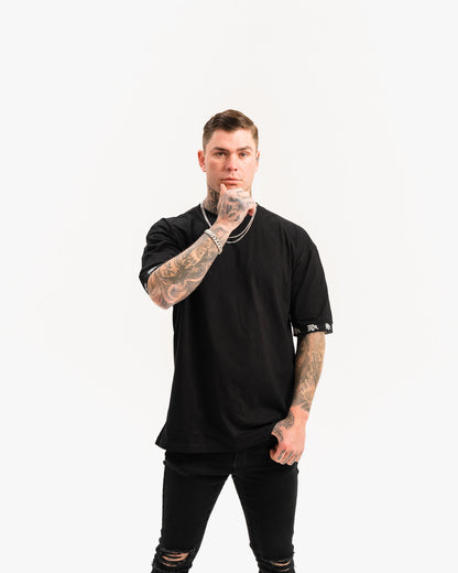 male model with tattoos and chains wearing black t-shirt and black ripped jeans, white background