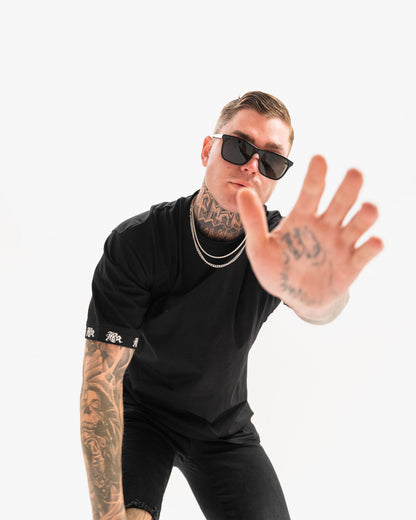 male model with tattoos, chains, sunglasses wearing black t-shirt and black ripped jeans, white background