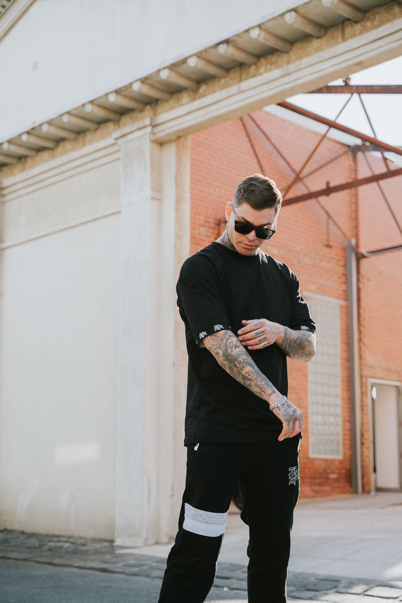 male model with tattoos, sunglasses and chains wearing black t-shirt and black pants, urban background