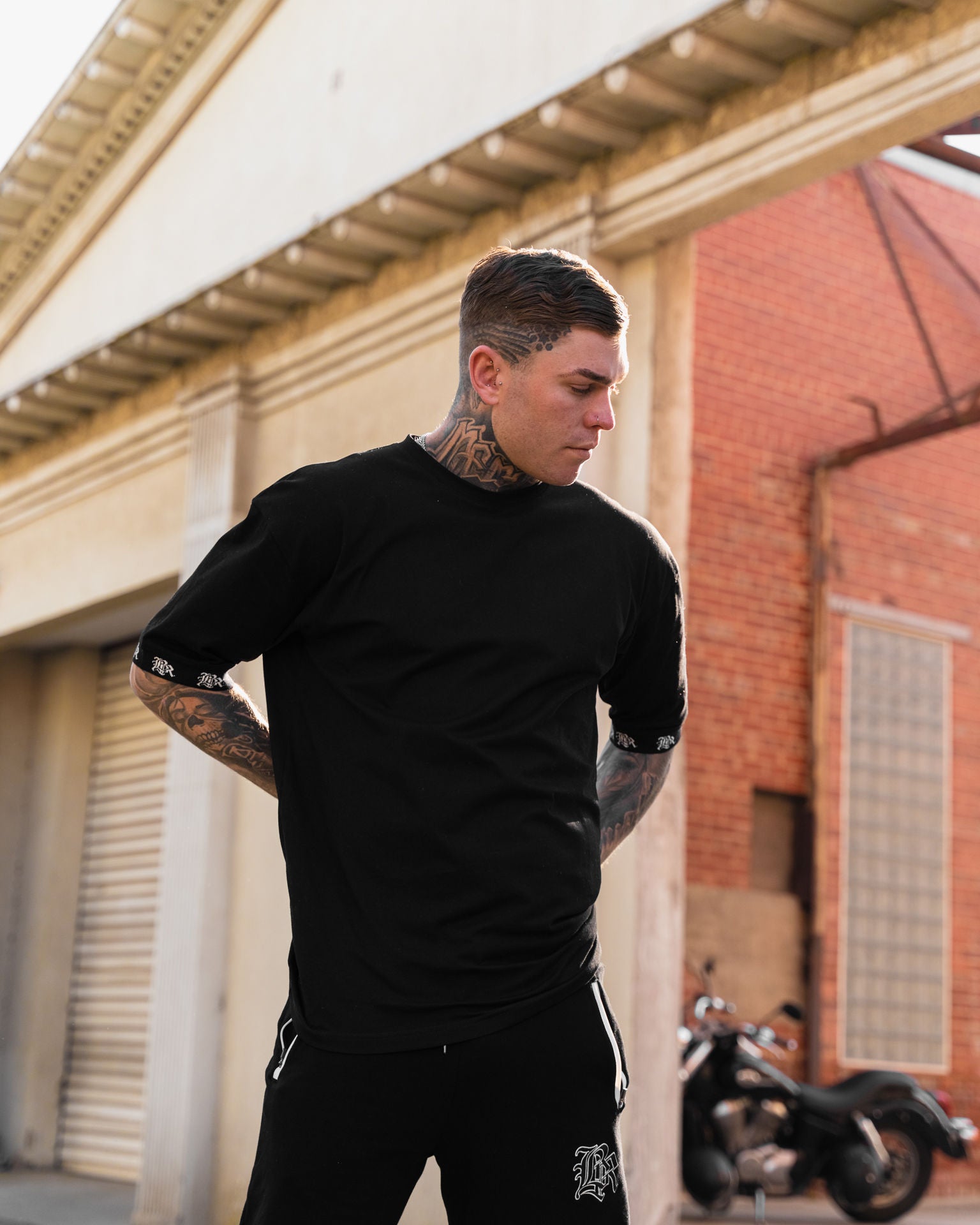 male model with tattoos, sunglasses and chains wearing black t-shirt and black pants, urban background