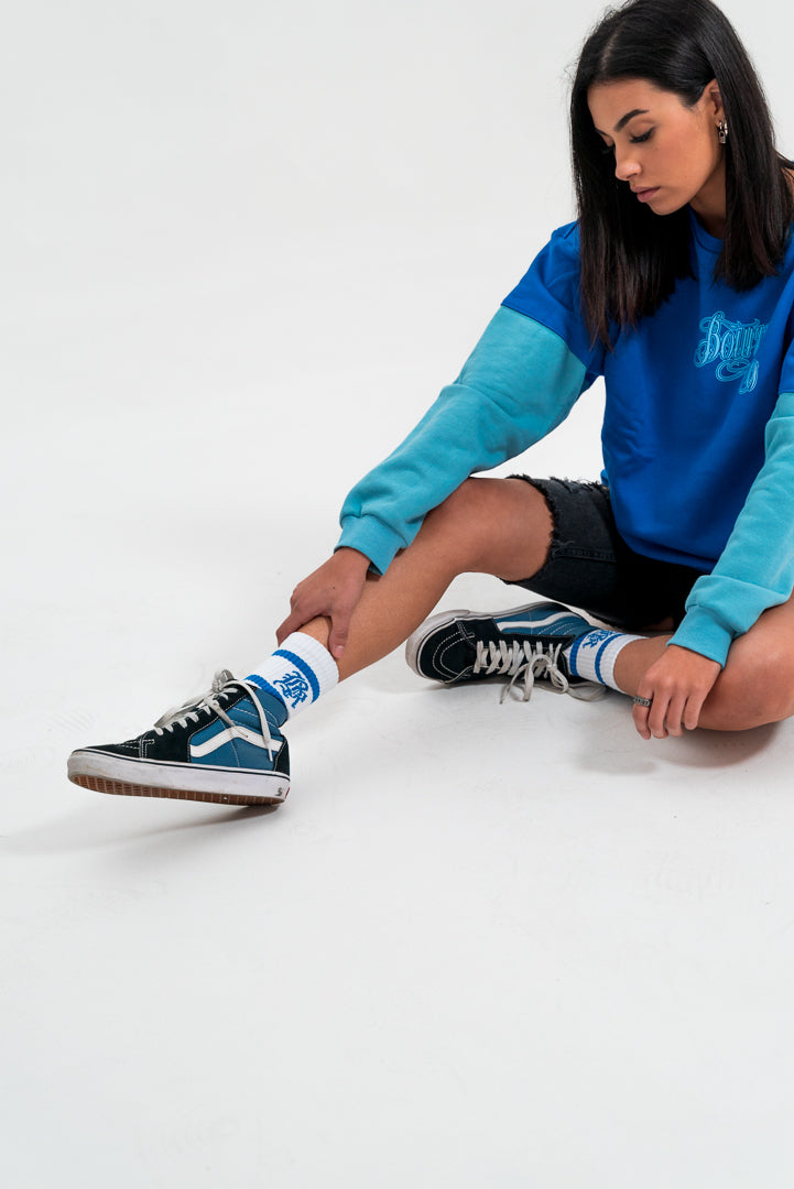 female model looking away, wearing a blue and teal crewneck hoodie and black bottoms, white background