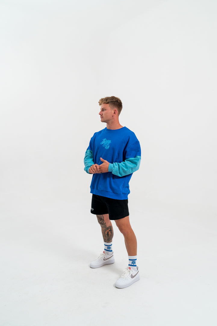 male model looking away, wearing a blue and teal crewneck hoodie and black bottoms, white background