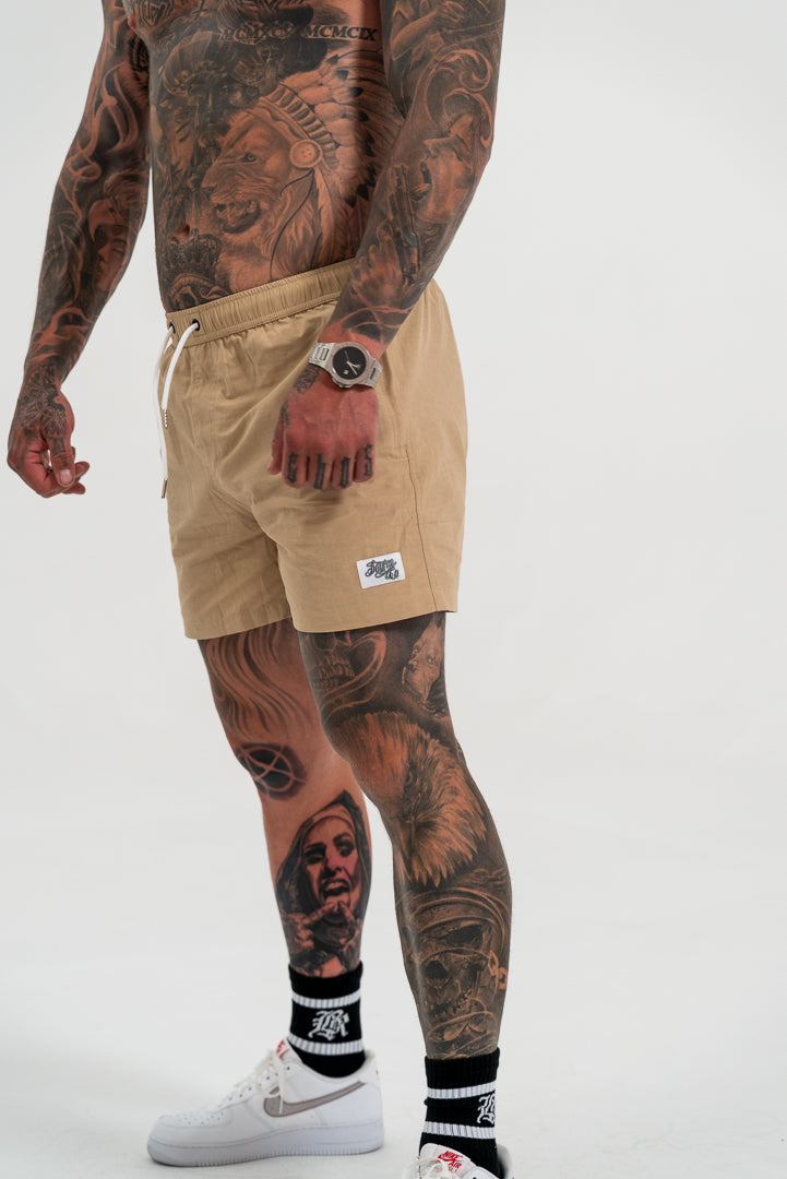 male model with tattoos, wearing tan swim shorts and white shoes, white background