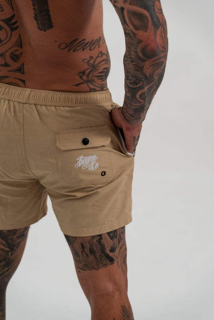 male model with tattoos, wearing tan swim shorts and white shoes, white background