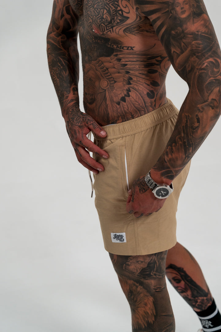 male model with tattoos, wearing tan swim shorts and white shoes, white background