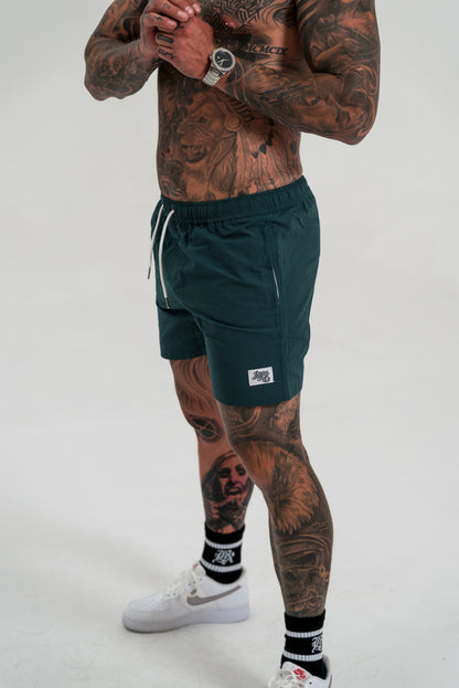 male model with tattoos wearing green short, white shoes and black socks, white background