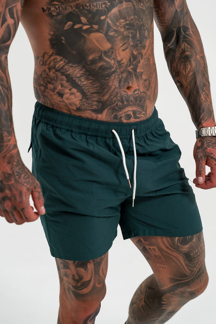 male model with tattoos wearing green short, white shoes and black socks, white background