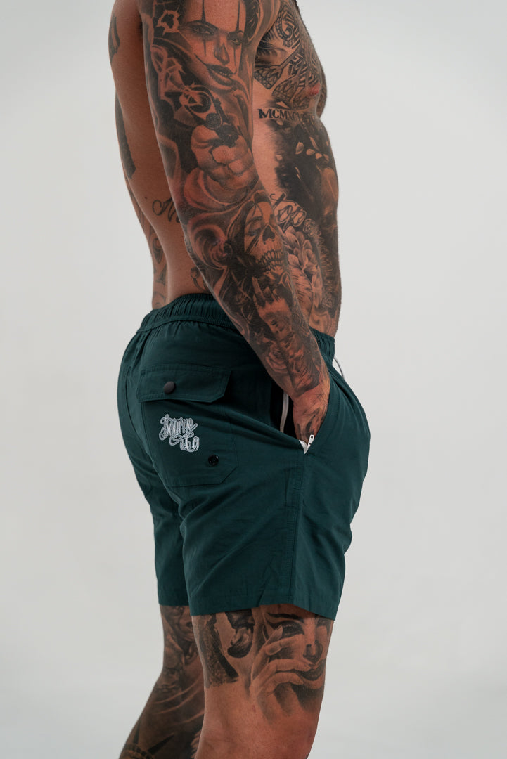 male model with tattoos wearing green short, white shoes and black socks, white background