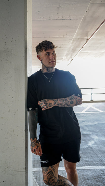 male model with tattoos, sunglasses and chains wearing black t-shirt and black pants, urban background