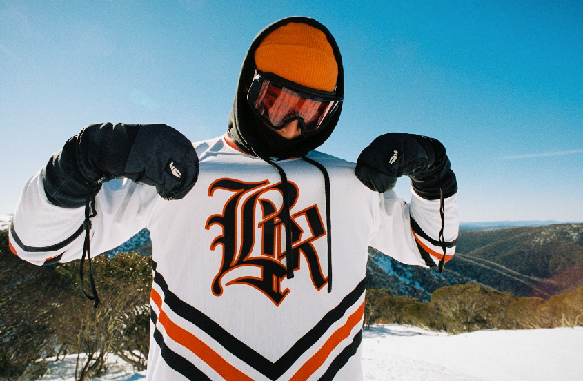 snowboarder showing off snow jersey, orange beanie, white snow jersey, mountains in the background