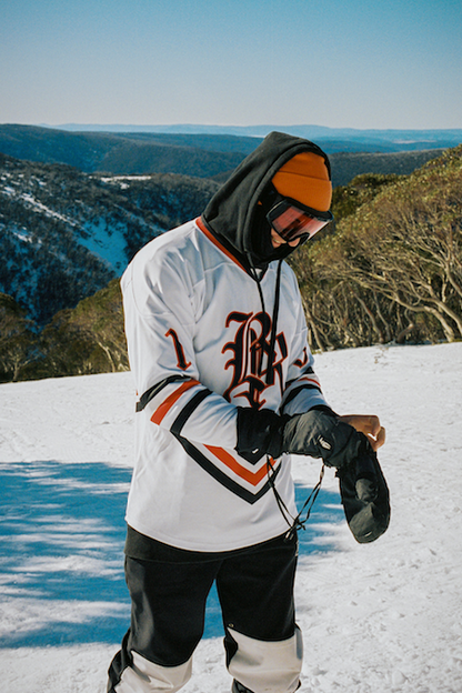 snowboarder putting on gloves, orange beanie, white snow jersey, mountains in the background